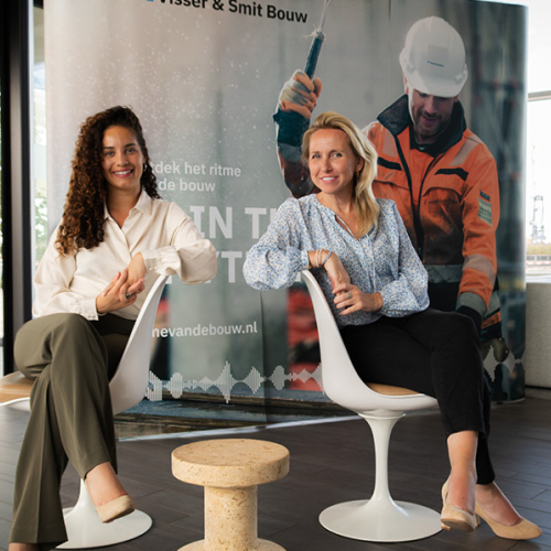 Djisri en Anita op zitten op twee stoelen in een lichte ruimte. Op de achtergrond een man met een oranje jas aan en helm op